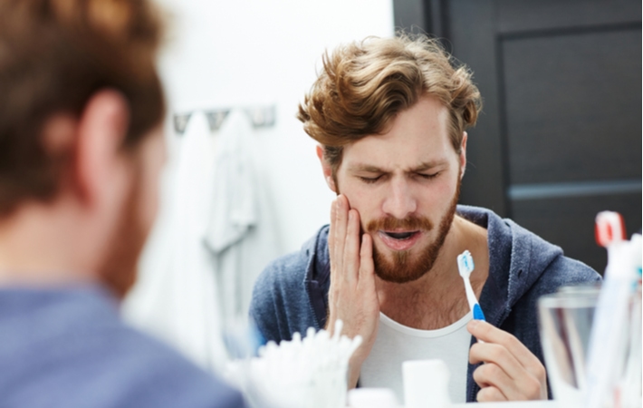 A man holding his cheek in pain while he brushes his teeth