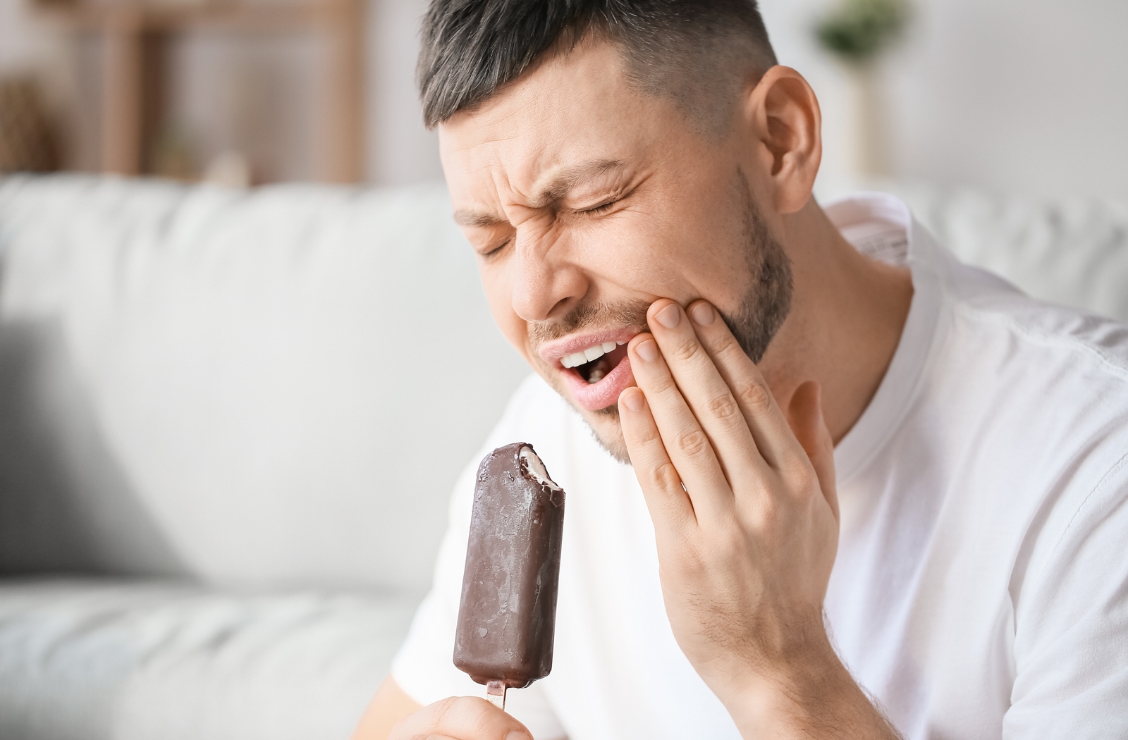 A man wincing in pain while holding his cheek after biting into a popsicle, as his teeth are sensitive to the cold