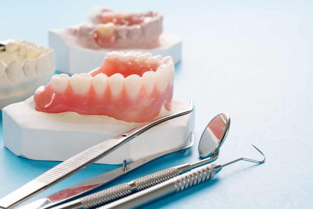 Dentures sitting on a white tray with models of teeth behind and dental tools in front.