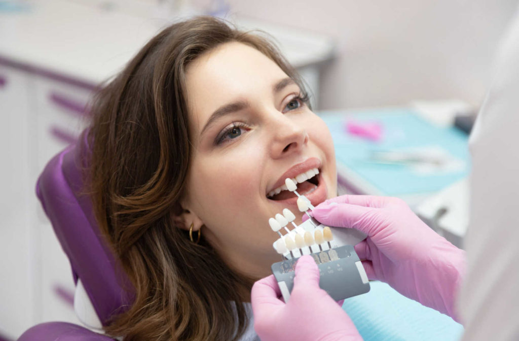 A woman sitting on a dental chair with an open mouth showing her teeth and a hand of a dentist holding a tooth color chart and doing a color selection on the patient's teeth.