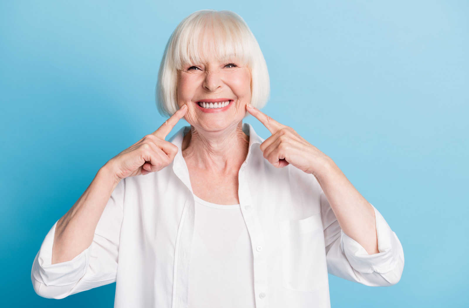 A lady in a white shirt with her two index finger pointing at her dentures with a big smile on her face.