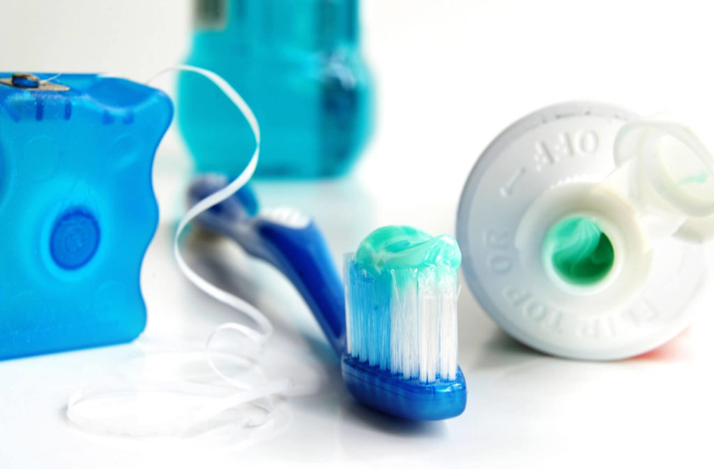 A full dental care bundle with toothbrush with toothpaste on it, floss, an open tube of toothpaste and a bottle of mouthwash in the background.