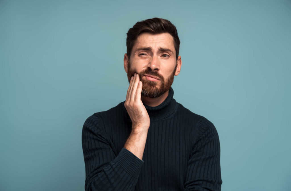 A man wearing a black turtleneck against a blue background holds his right hand to his cheek, indicating a toothache.