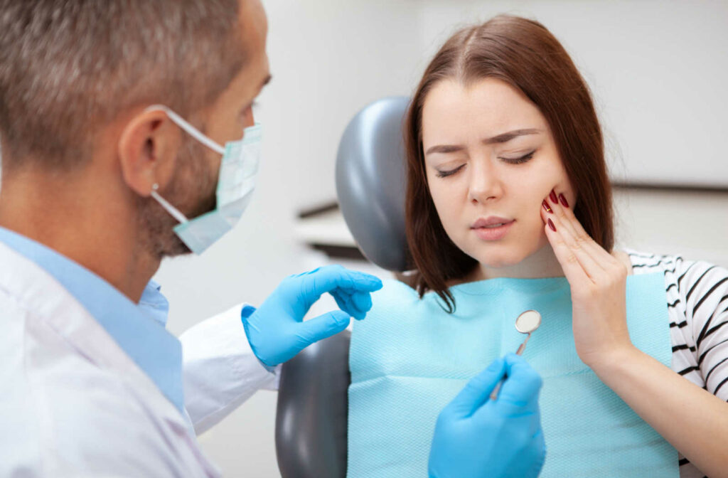 A young woman holding her left cheek with her left hand while her dentist prepares to examine her tooth.
