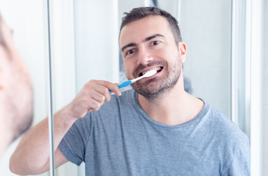 A man wearing a blue shirt with a toothbrush in his mouth smiling as he cleans his teeth.