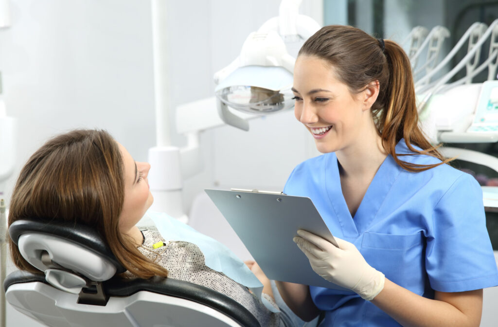 A female dentist in blue scrubs holding a clipboard and smiling at her patient in the dental chair.