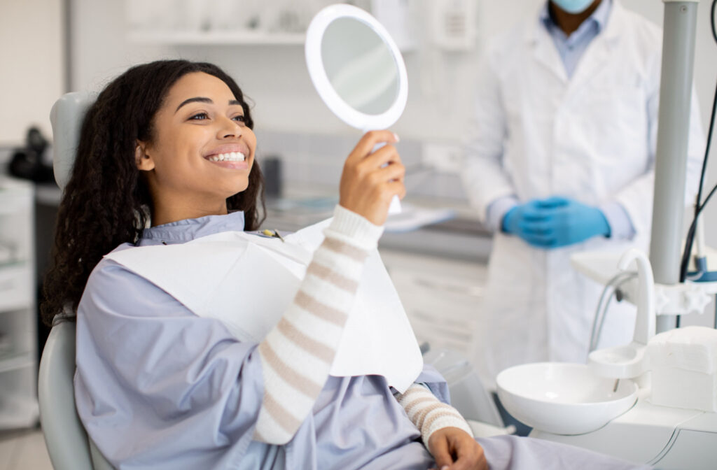 A woman sitting in a dentist's chair, holding up a mirror and smiling with her dentist in the background