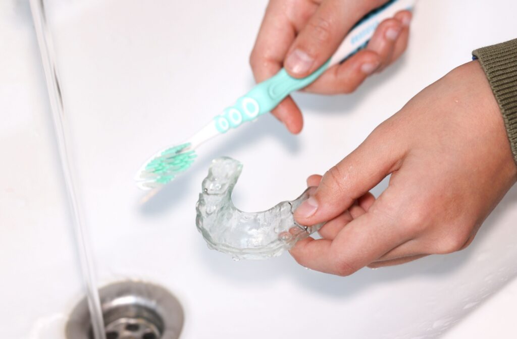 A person holding a clear dental aligner in a sink to clean it with a tooth brush and running water.